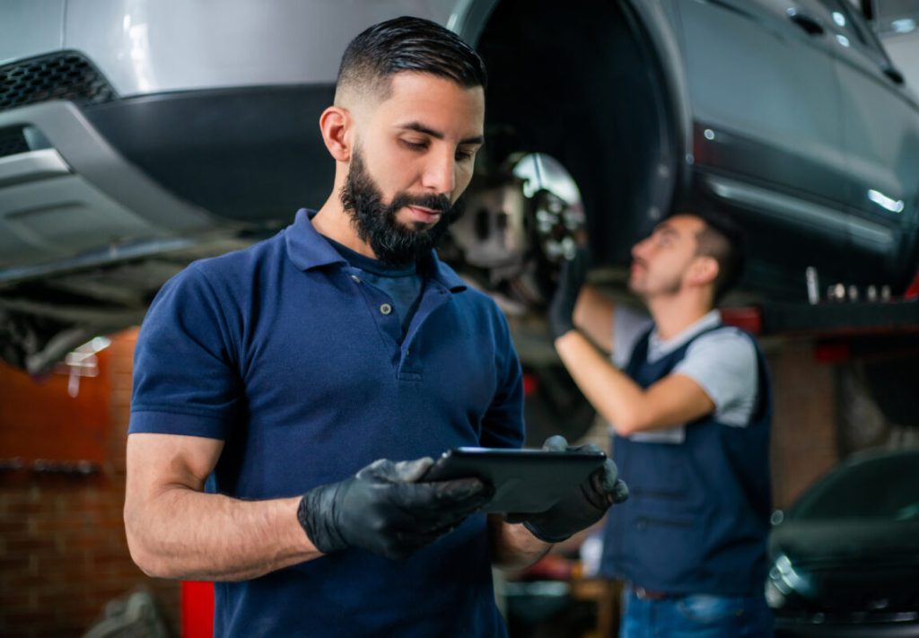 Supervisor at a car workshop checking tablet while mechanic works at background on a car
